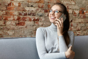 Pretty young fair-haired female with pony tail wearing big glasses and casual sweater sitting at cozy sofa communicating over mobile phone having charming smile looking aside isolated over brick wall
